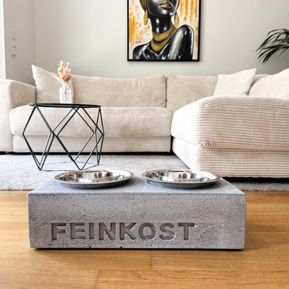 Napfhalter aus grauem Beton mit personalisiertem Namen und zwei Edelstahlnäpfen, frontal im Wohnraum